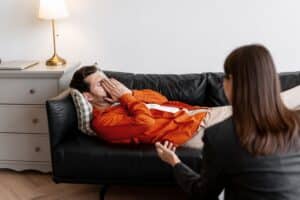 Man lying on couch with hand on forehead appearing distressed during therapy session with counselor sitting nearby holding clipboard