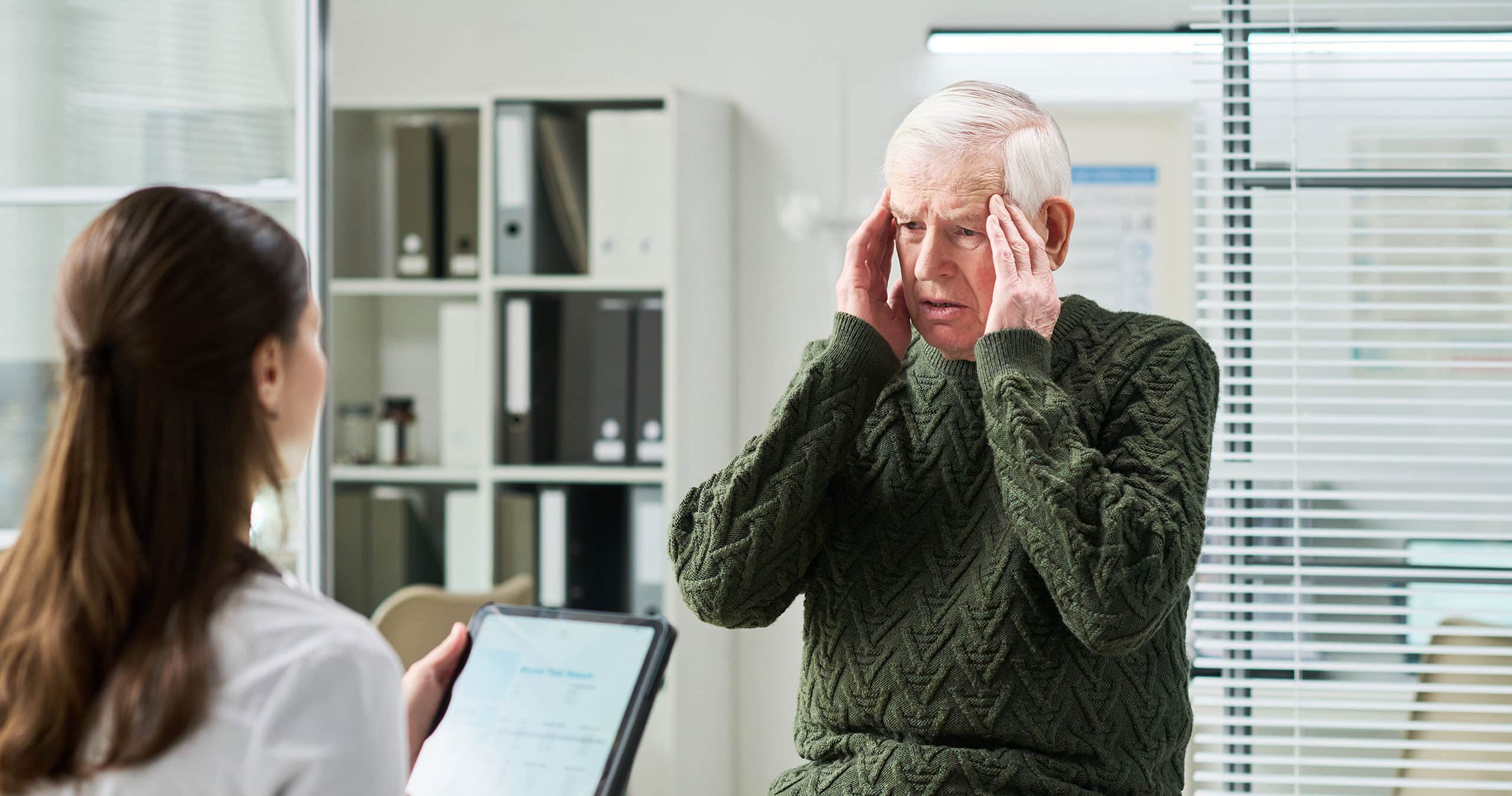 A senior man on examination table holding his head while female doctor with clipboard listens to him during medical consultation.